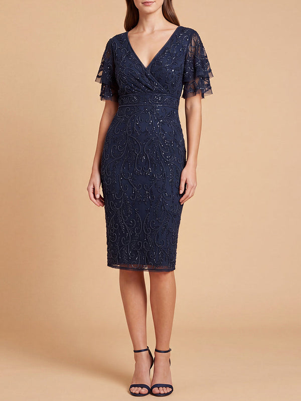 A woman modeling a navy blue beaded midi dress with short flutter sleeves, a V-neckline, and a fitted silhouette, standing against a solid beige studio background.