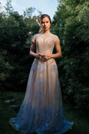A woman wearing a blue dress stands in a grassy area, surrounded by nature, with a serene expression.
