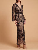 A woman models a long-sleeve black lace maxi dress with a V-neckline and sheer fishnet overlay, set against a solid warm beige studio background.