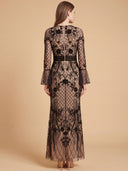 A woman viewed from the back wearing a long black lace maxi dress with floral embroidery and sheer mesh panels, standing against a plain beige studio background.