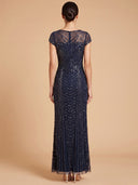A woman viewed from the back wearing a floor-length navy evening gown with sheer cap sleeves, a sheer upper back panel, and dense beaded geometric patterns running down the skirt, against a plain beige studio backdrop.