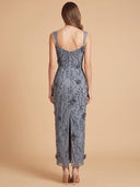 A woman viewed from the back wearing a sleeveless, midi-length gray dress with floral embroidery and a low open back, standing against a plain beige studio background.