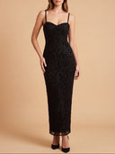 A woman modeling a black spaghetti-strap evening gown with intricate beaded and sequined vertical detailing, set against a solid warm beige studio background.