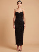 A woman modeling a black spaghetti-strap, floor-length gown covered in intricate beadwork and sequins, set against a solid warm beige studio background.