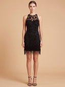 A model wearing a sleeveless black lace and bead fringe mini dress with a high neckline, standing against a solid beige studio background.