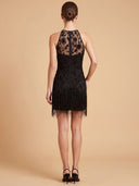 A woman shown from the back wearing a sleeveless black dress with floral lace panels and fringe, photographed against a plain beige studio background.