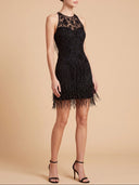 A woman models a sleeveless black mini dress with a sheer lace bodice, beaded texture, and a fringe hem, paired with black high-heeled sandals against a solid beige studio background.