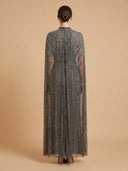 A woman viewed from the back wearing a floor-length sheer gray cape-style gown with dense beaded embroidery and a central back opening, set against a solid warm beige studio background.