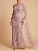 A woman models a floor-length, one-shoulder evening gown in a dusty rose pink color with intricate silver sequin embroidery and a matching sheer draped cape. The background is a solid warm beige studio backdrop.