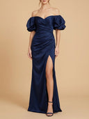 A woman modeling an off-the-shoulder navy blue satin gown with puffed sleeves, a sweetheart neckline, ruched draping, and a high thigh slit, set against a solid beige studio background.