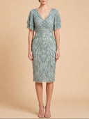 A woman modeling a sage green beaded midi dress with a V-neckline, short flutter sleeves, and a fitted silhouette against a solid beige background.