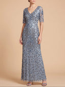 A woman modeling a floor-length, short-sleeve V-neck gown covered in shimmering silver and blue sequins and beadwork, photographed against a plain warm beige studio background.