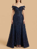 A woman modeling an off-the-shoulder navy blue lace and sequin floor-length gown against a solid beige studio background.