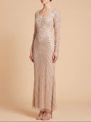 A woman models a floor-length champagne beige gown with intricate beaded sunburst patterns and long sleeves, set against a solid warm beige studio background.