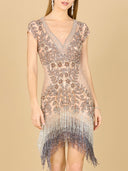 A woman models the Lara 29609 Beaded Fringe Short Cocktail Dress, featuring a V-neck, silver and blush pink beaded floral patterns, and shimmering fringe. She pairs it with long earrings and a bracelet against a beige background.