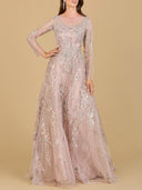 A woman wears the Lara 29618 Beaded Long Sleeve A-line Dress—a blush pink, modest gown with sheer long sleeves, silver embroidery, a fitted bodice, and flowing floor-length skirt—standing against a beige background.