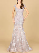 A woman wears the Lara 29642 Cap Sleeve Mermaid Gown, a fitted, floor-length lavender dress with floral lace embroidery, cap sleeves, sweetheart neckline, and flared semi-sheer skirt. She stands against a beige background.