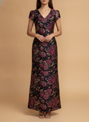 A woman modeling a floor-length V-neck maxi dress with short cap sleeves, featuring a dark floral jacquard pattern in shades of purple, burgundy, and beige, set against a solid beige studio background.