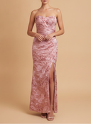 A woman models a strapless, floor-length pink gown with a tonal floral jacquard pattern and a high front slit, set against a solid beige studio background.