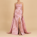 A woman models a floor-length pink gown with a textured, shimmering jacquard pattern, spaghetti straps, a fitted bodice, and a high thigh slit. She is standing against a solid beige studio background.