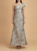A woman modeling a floor-length one-shoulder evening gown with an intricate floral jacquard pattern in muted gray, beige, and gold tones, set against a solid warm beige studio background.