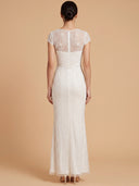A woman viewed from the back wearing a fitted white lace floor-length gown with short cap sleeves and a sheer lace back panel, standing against a warm beige studio background.