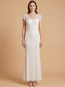 A woman modeling a floor-length white lace gown with cap sleeves and a V-neckline, standing against a warm beige studio backdrop.