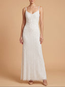 A woman modeling a floor-length white sleeveless gown with thin straps, a V-neckline, and textured beaded detailing, standing against a warm beige studio backdrop.