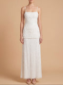 A woman modeling a white sleeveless maxi dress with thin straps, a fitted textured bodice, and a flowing pleated skirt, photographed against a warm beige studio background.