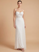 A woman modeling a sleeveless white maxi gown with thin straps, a fitted embellished bodice, and a flowing tulle skirt against a warm beige studio background.