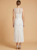 A woman viewed from the back wearing a sleeveless white lace midi dress with floral embroidery and sheer upper back panels, paired with metallic high heels, against a warm beige studio backdrop.