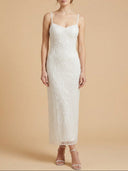 A woman modeling a sleeveless, floor-length white lace dress with thin straps against a warm beige studio background.