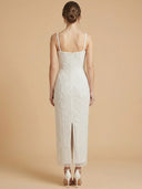 A woman viewed from the back wearing a sleeveless ivory lace midi dress with thin straps, a fitted silhouette, and a front slit, standing against a warm beige studio background.