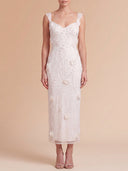 A woman modeling a sleeveless white lace midi dress with a sweetheart neckline, thin shoulder straps, and 3D floral appliques, set against a solid beige studio background.