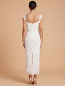 A woman viewed from the back wearing a fitted white lace midi dress with cap sleeves, a low open back, and a front slit, standing against a warm beige studio backdrop.