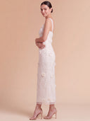 A woman modeling a sleeveless white lace midi dress with 3D floral applique, shown from a side-back angle against a solid beige studio backdrop.