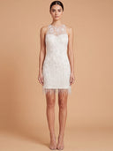 A woman modeling a sleeveless white lace mini dress with fringe trim against a solid beige studio background.