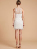 A woman shown from the back wearing a short white lace and fringe dress against a warm beige studio background.