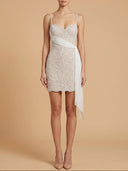 A woman modeling a short beaded mini dress with thin straps, a sweetheart neckline, and a draped side detail, photographed against a plain warm beige studio background.