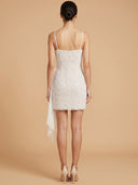 A woman shown from the back wearing a short white beaded mini dress with thin straps and a draped side detail, photographed against a warm beige studio background.