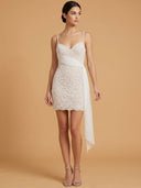 A woman models a short white beaded mini dress with thin spaghetti straps, a sweetheart neckline, a draped waist, and a long flowing side skirt, photographed against a solid warm beige studio background.