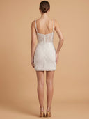 A woman shown from the back wearing a fitted mini dress with thin straps, lace detailing at the upper back, and a pleated textured skirt, photographed against a warm beige studio background.
