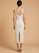A woman shown from the back wearing a sleeveless ivory lace midi dress with a deep V back and front slit, standing against a warm beige studio background.