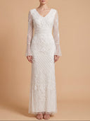A woman modeling a long-sleeve white lace maxi dress with a V-neckline and fitted silhouette, standing against a solid warm beige studio background.
