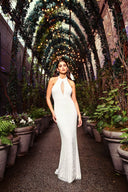 A woman in a flowing white gown stands gracefully under a floral arbor, surrounded by greenery.
