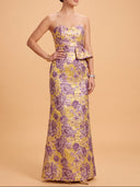 A woman modeling a strapless floor-length gown with a purple and gold floral jacquard pattern against a solid peach studio background.
