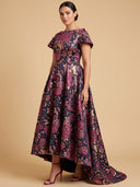A woman models a floor-length floral gown with short flutter sleeves, a fitted waist, and a high-low skirt with a slight train, in a dark purple and pink floral jacquard pattern against a solid beige studio background.