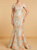 A woman models a floor-length off-the-shoulder gown with a square neckline, fitted bodice, and flared mermaid hem. The dress has an allover floral pattern in peach-orange and sage green on a light beige base, set against a solid warm beige studio backdrop.