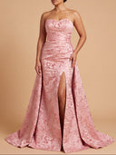 A woman models a strapless pink gown with a sweetheart neckline, textured floral jacquard pattern, and a high thigh slit, set against a solid beige studio background.