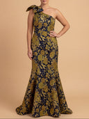 A woman modeling a one-shoulder floor-length gown with a large bow detail on the shoulder and an allover floral jacquard pattern in olive green and navy blue, set against a solid beige studio background.